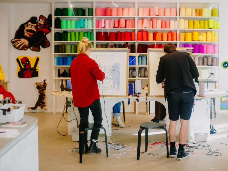 Two people are creating designs on canvas using tufting guns and yarn